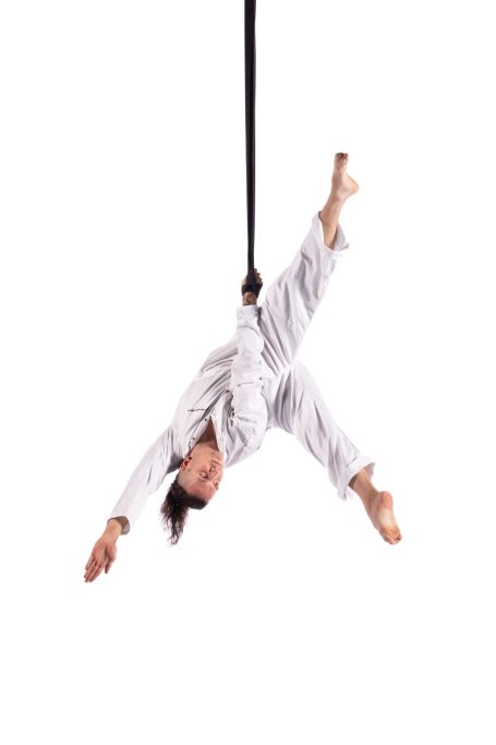 A performer in a white boiler suit hangs upside down from one arm.