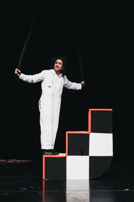 A performer in a white boiler suit stands at the base of a checkerboard staircase.