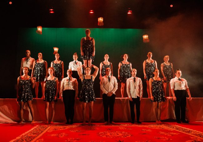 Many performers all smartly dressed stand in front of and on top of a long dinner table. A single performer stands on another's shoulders.