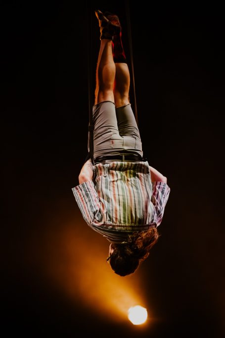 A performer in a striped shirt is inverted with his hands in his pockets suspended by aerial straps.