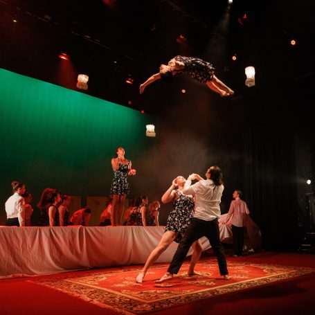 A performer in a blue dress does a flip high above two smartly dressed performers underneath, prepared to catch her.