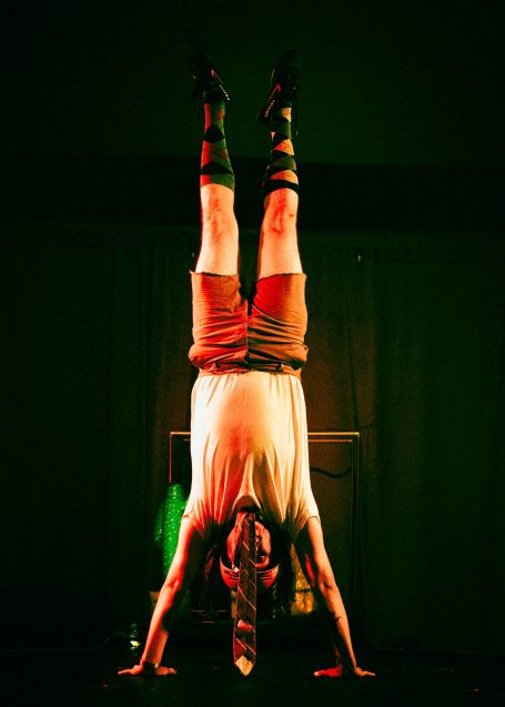 A performer in argyle socks and a tie holds a handstand while looking out at the audience.