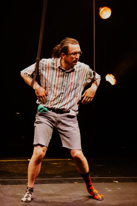A performer in a striped shirt stands with his arms suspended in straps.