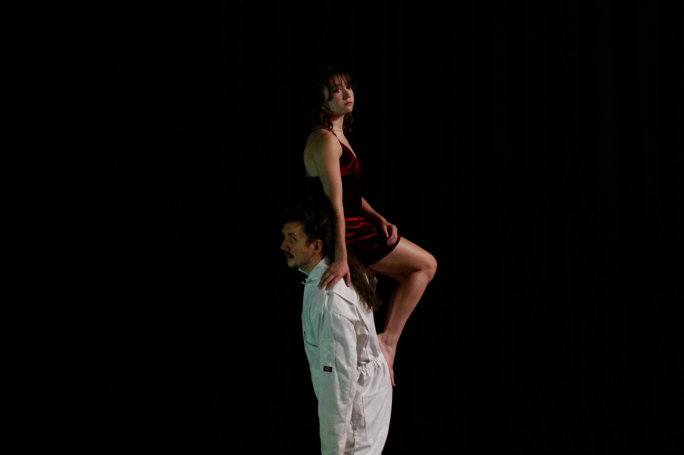 One performer in a boiler suit stands looking into the distance, a second performer in a red dress sits on his shoulder.