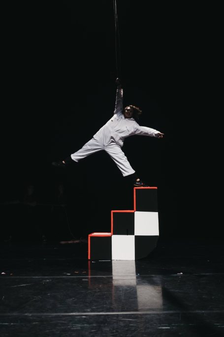 A performer in a white boiler suit hangs from straps with one foot in the air and the other on top a checkerboard staircase. 