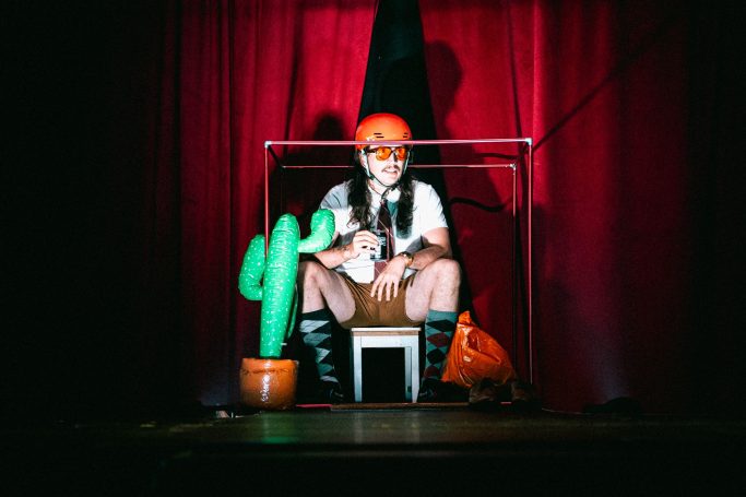 A performer in an orange helmet sits on a tiny chair with an inflatable cactus.