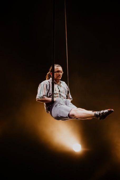 A performer in a striped shirt is suspended in a seated position hands in pockets, from aerial straps.